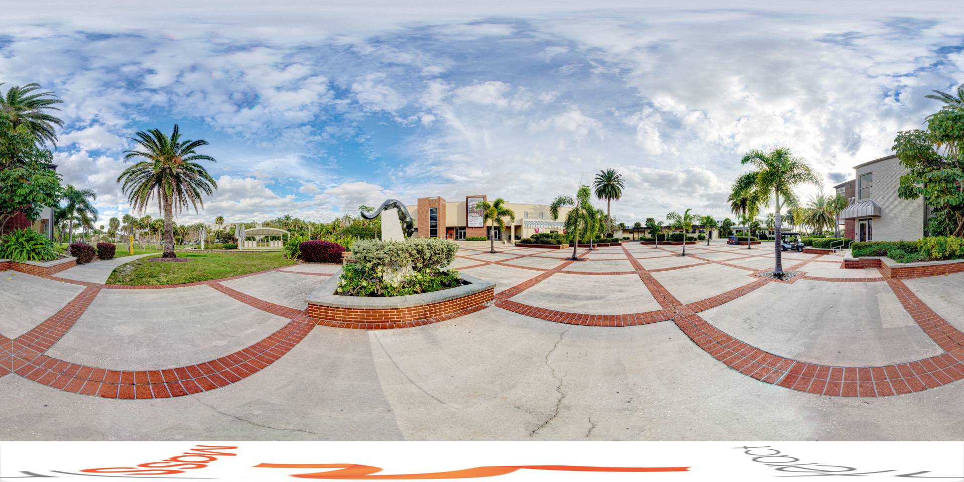 Panoramic view of a tropical campus courtyard with palm trees, brick pathways, and modern buildings under a cloudy sky
